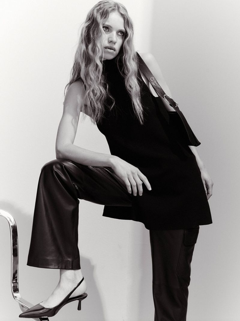 A woman models black sleeveless top with matching purse and dark leather pants, posing indoors against a neutral backdrop. Black heels visible.