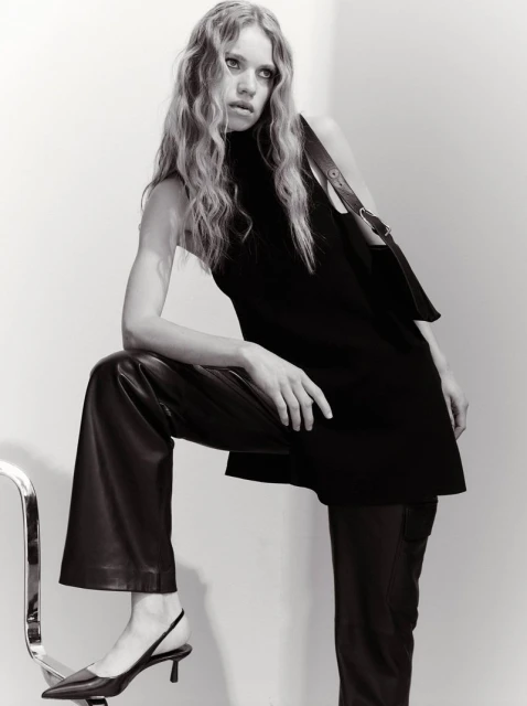 A woman models black sleeveless top with matching purse and dark leather pants, posing indoors against a neutral backdrop. Black heels visible.