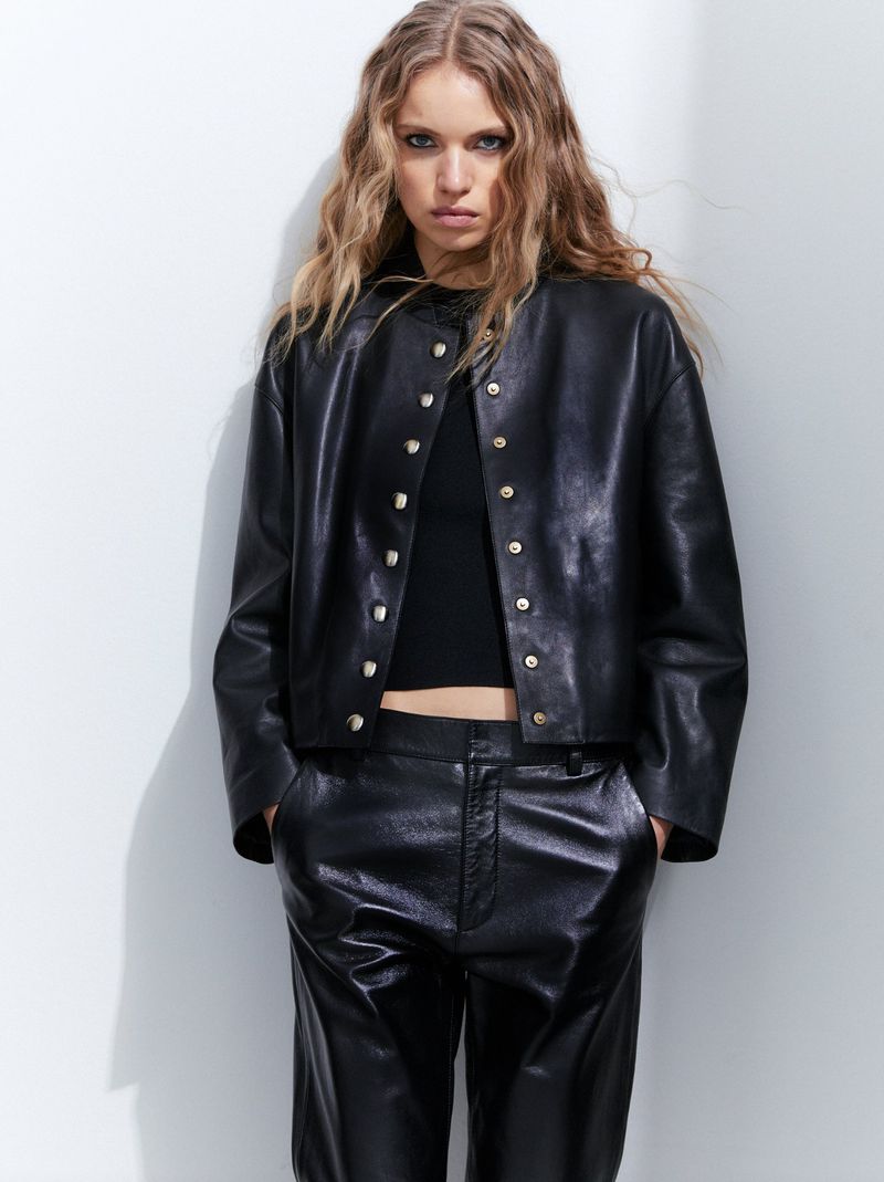 Blonde woman in black leather jacket and pants poses in front of a white wall for a fashion editorial shot in a photo studio.