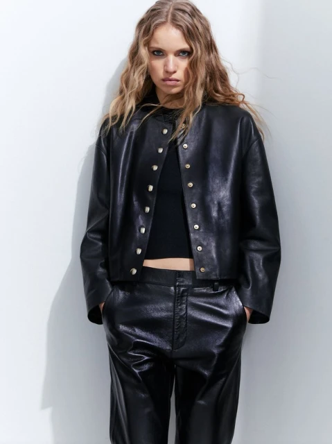 Blonde woman in black leather jacket and pants poses in front of a white wall for a fashion editorial shot in a photo studio.