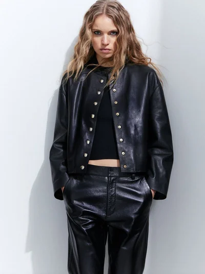 Sabine Glud posing in a black leather jacket with metallic snap buttons and matching leather trousers against a white studio wall.