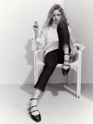 Model Sabine Glud seated in a white chair wearing a white top, dark cropped pants, and patent multi-strap flats in black and white.