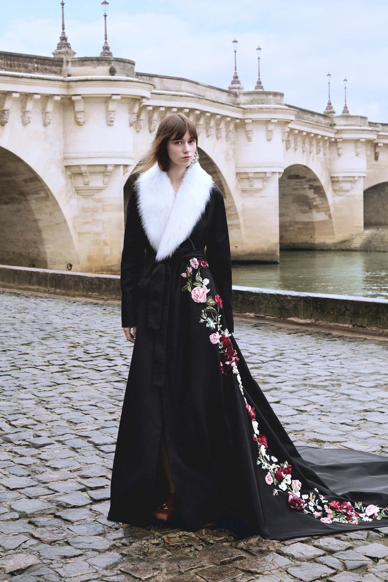 A woman in a long black coat with floral embroidery and a white fur collar stands on a stone walkway next to a river and bridge.