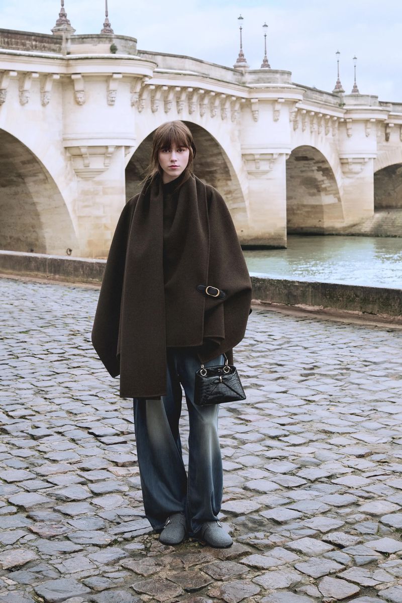 A fashionable adult female stands on a cobblestone walkway wearing a brown coat, blue jeans, a black purse, and gray shoes near a bridge.