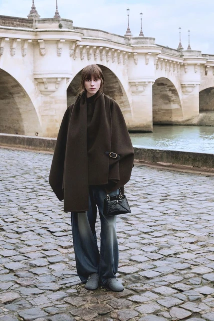 A fashionable adult female stands on a cobblestone walkway wearing a brown coat, blue jeans, a black purse, and gray shoes near a bridge.