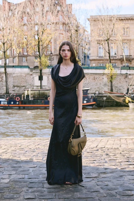 Woman in a long black dress standing in front of a river in an urban setting, holding a purse. Buildings and trees in background.