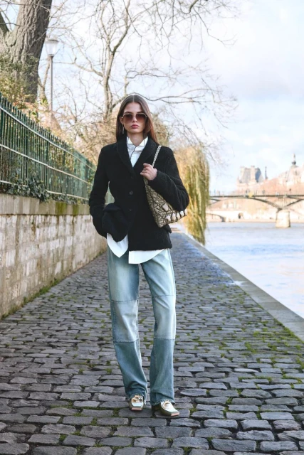 A woman in a blazer, shirt, and jeans stands on a cobblestone path with a river and buildings in the background in an outdoor setting.
