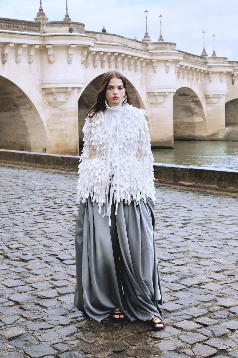 Fashion model in a white fringed jacket and grey wide-leg pants posing on a cobblestone bridge with stone arches in the background.
