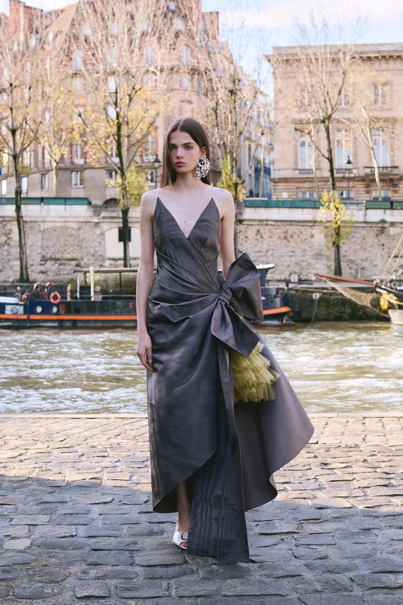 A woman stands on a stone walkway near a river and buildings. She wears a long grey dress with a large bow and statement earrings.