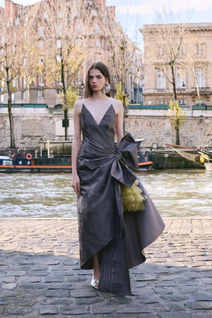A woman stands on a stone walkway near a river and buildings. She wears a long grey dress with a large bow and statement earrings.