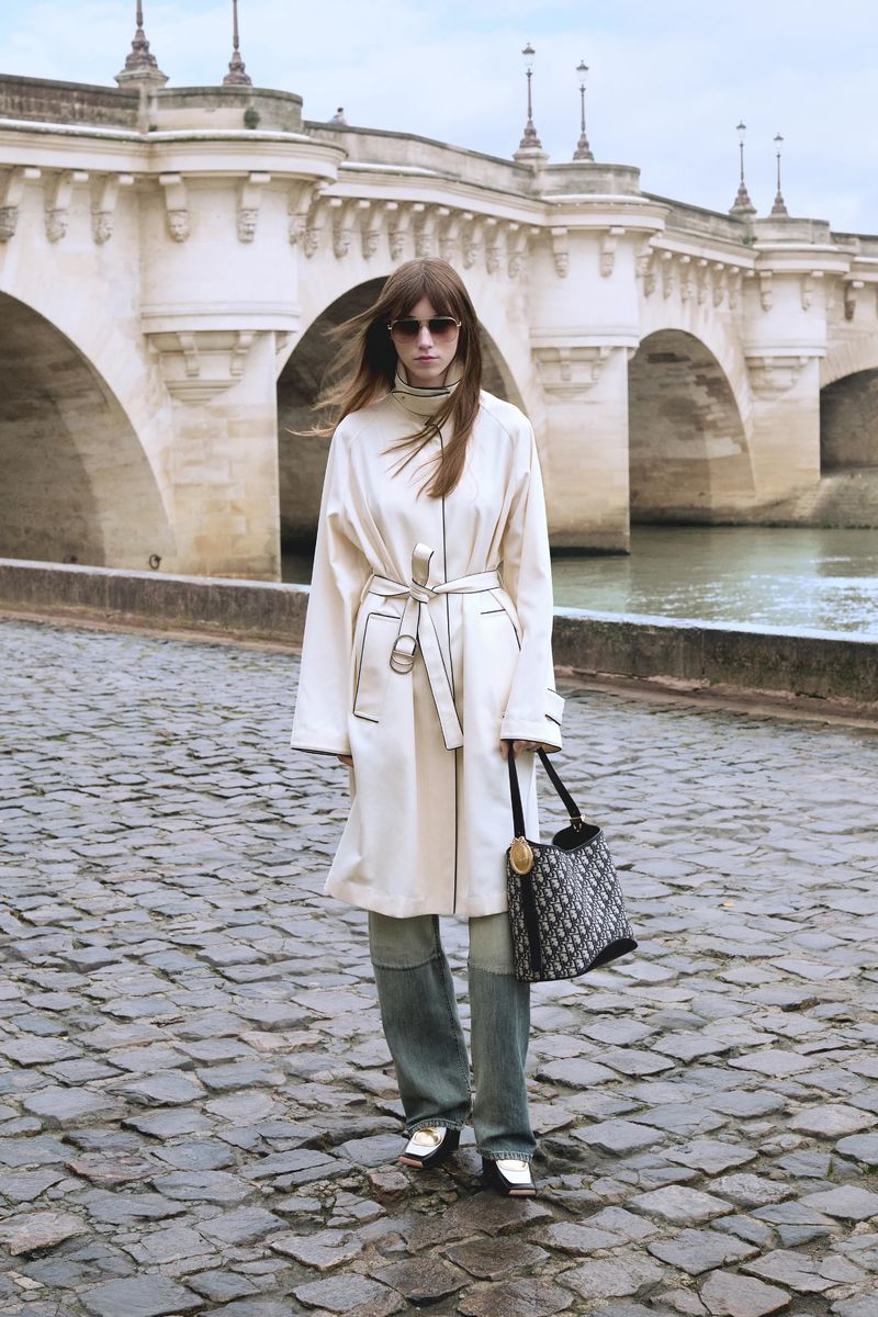 Woman in cream coat and jeans stands on cobblestone street with handbag, Paris bridge in background, fashion photography.