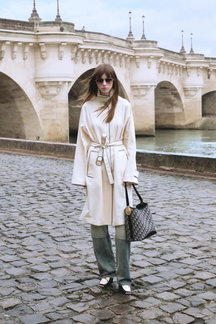 Woman in cream coat and jeans stands on cobblestone street with handbag, Paris bridge in background, fashion photography.
