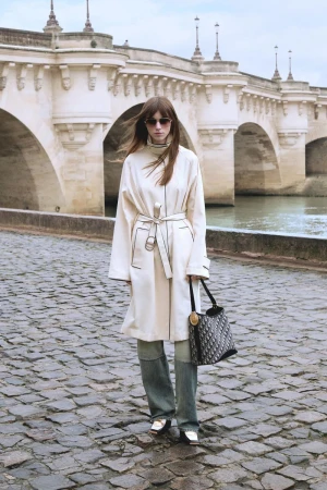 Woman in cream coat and jeans stands on cobblestone street with handbag, Paris bridge in background, fashion photography.