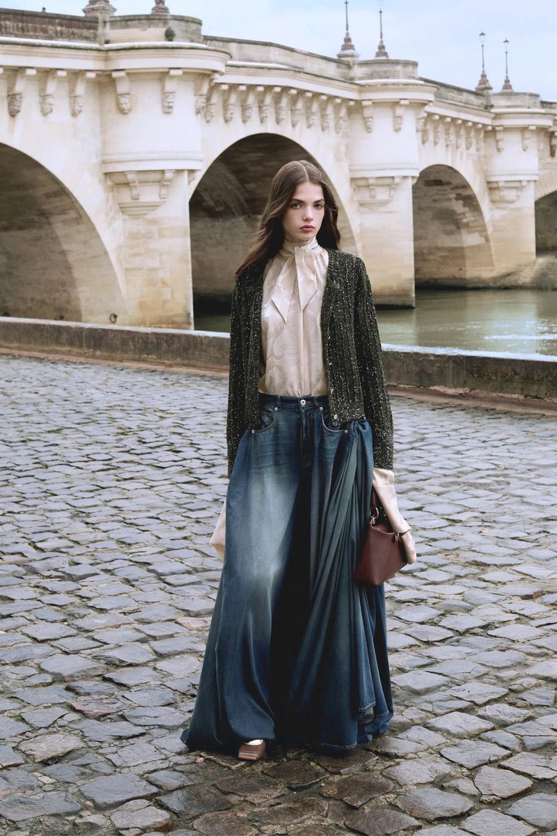Fashion shot of a woman in a long denim skirt, blouse and sweater on a cobblestone bridge in Paris with the Seine in the background.