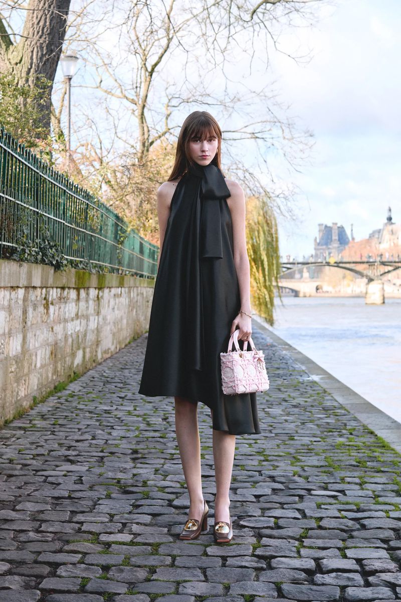 Woman poses on cobblestone path in a black halter dress with bow, holding a pink purse, city river and buildings in background.