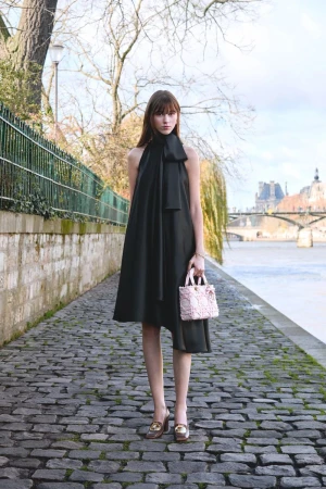 Woman poses on cobblestone path in a black halter dress with bow, holding a pink purse, city river and buildings in background.