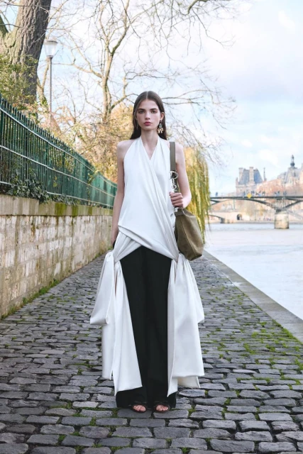 Fashion model in a draped white top and black pants poses on a cobblestone path near the Seine River in Paris on an overcast day.