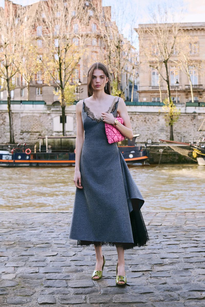 A fashionable woman in a grey dress stands on a stone walkway by a river in an urban setting, holding a pink clutch, with buildings visible.