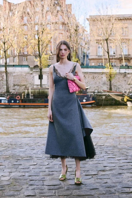 A fashionable woman in a grey dress stands on a stone walkway by a river in an urban setting, holding a pink clutch, with buildings visible.