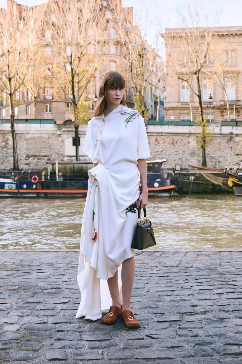 Fashionable woman in white dress stands near river with handbag and loafers; backdrop of Parisian buildings and boats on the water.