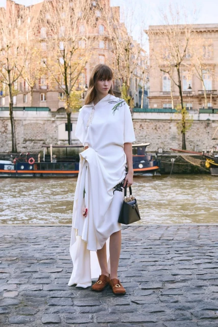 Fashionable woman in white dress stands near river with handbag and loafers; backdrop of Parisian buildings and boats on the water.