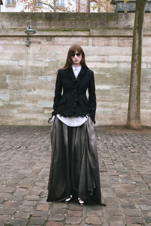Model standing on cobblestones wearing a black ruffled jacket, white shirt, and draped charcoal maxi skirt for Dior.