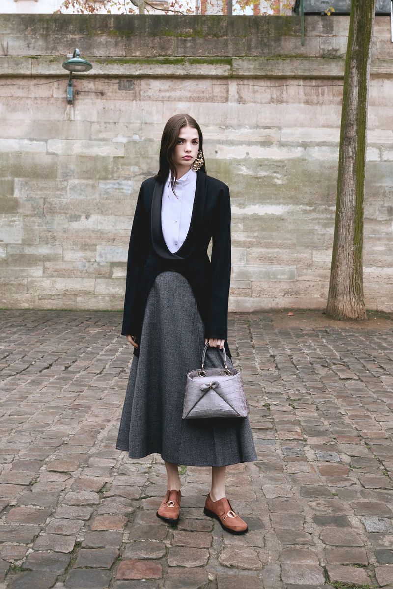 Full-shot of a woman in a black blazer, grey skirt, and brown shoes, holding a handbag, standing against a stone wall and cobblestone ground.
