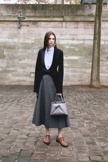 Full-shot of a woman in a black blazer, grey skirt, and brown shoes, holding a handbag, standing against a stone wall and cobblestone ground.