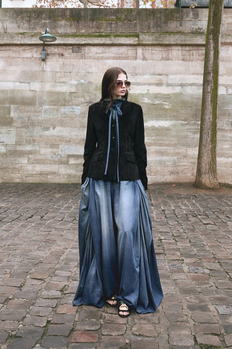 A female fashion model poses on a cobblestone street. She wears a black velvet jacket, a long blue-ombre skirt, sunglasses, and sandals.