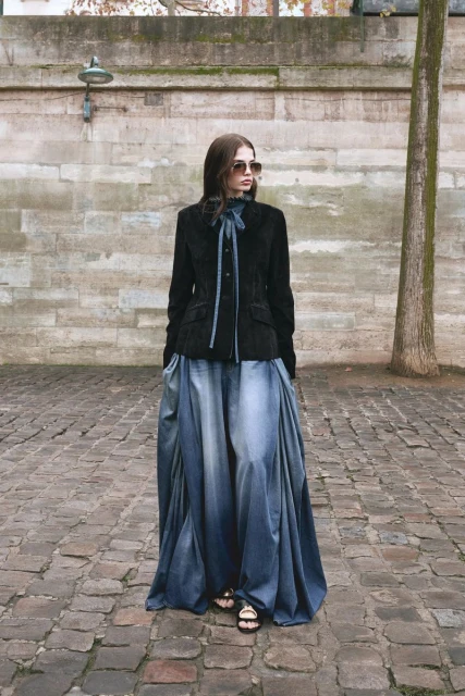 A female fashion model poses on a cobblestone street. She wears a black velvet jacket, a long blue-ombre skirt, sunglasses, and sandals.