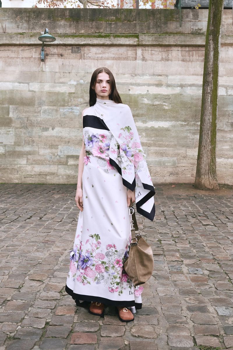 A woman in a floral gown and brown loafers stands on a cobblestone street in an urban setting, carrying a hobo bag in natural light.