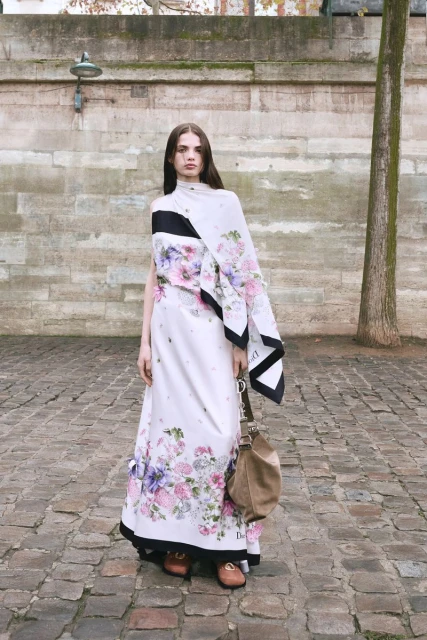 A woman in a floral gown and brown loafers stands on a cobblestone street in an urban setting, carrying a hobo bag in natural light.