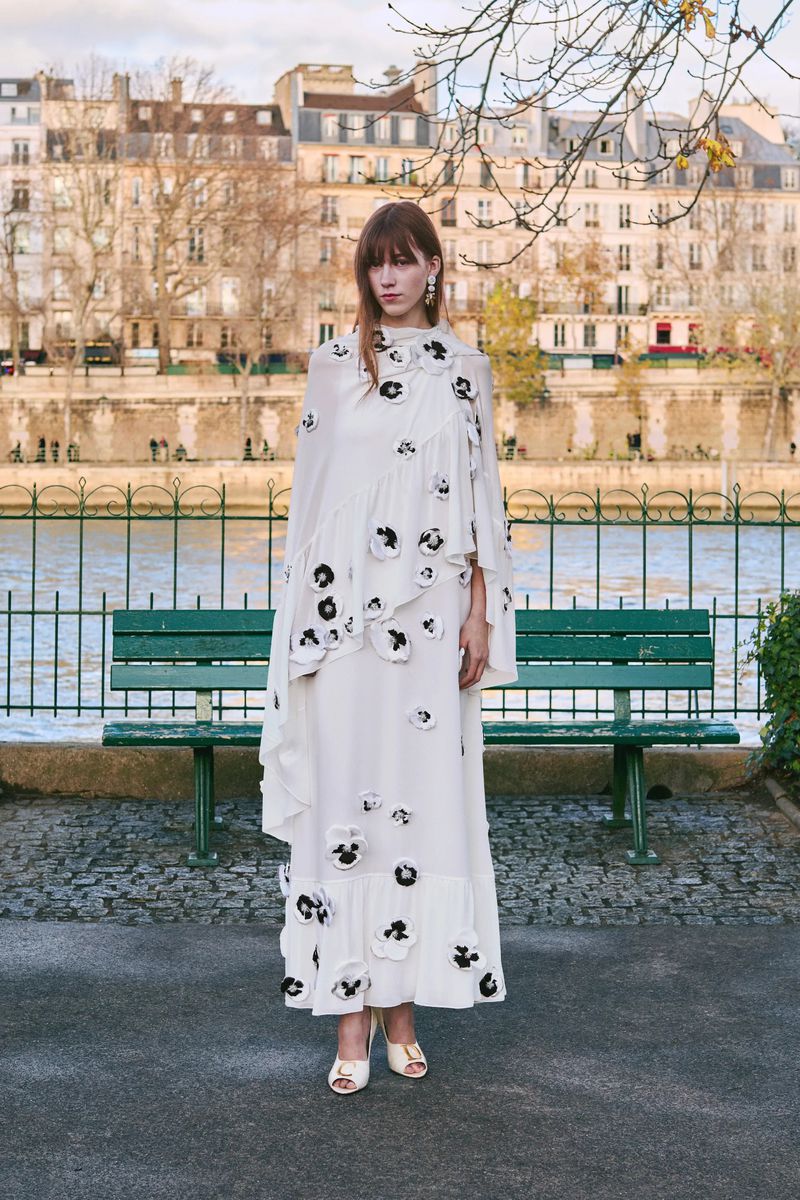 Woman in long white floral dress standing on a cobblestone path in front of a bench and river in an urban setting.
