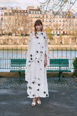 Woman in long white floral dress standing on a cobblestone path in front of a bench and river in an urban setting.