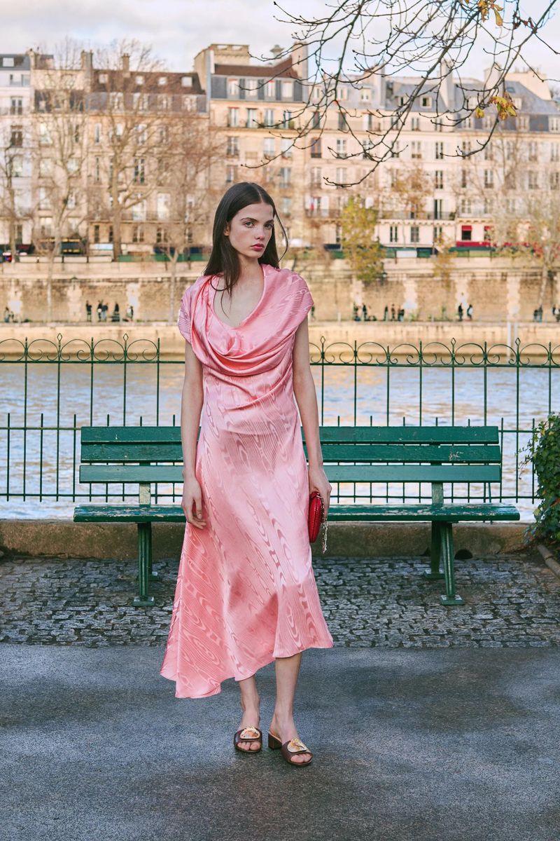 Brunette woman in pink dress and sandals stands outdoors on a sunny day in front of a river and European-style buildings.