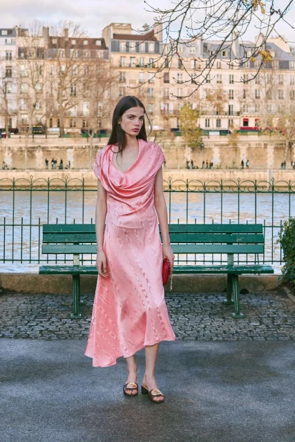 Brunette woman in pink dress and sandals stands outdoors on a sunny day in front of a river and European-style buildings.