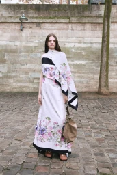 Model wearing a floral print dress and holding a taupe handbag standing outdoors on a cobblestone street