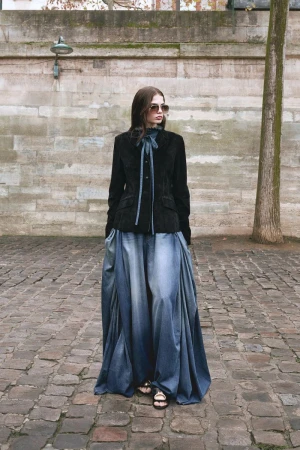 Model wearing a black tailored jacket, sunglasses, and an oversized flared denim maxi skirt on a cobblestone street.