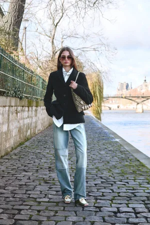 Woman wearing sunglasses, a black jacket, and light blue patchwork jeans standing on a cobblestone river walkway.