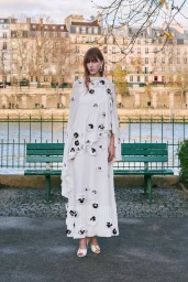 Model wearing a floor-length white floral appliqué cape dress standing outdoors in front of a green park bench and river.