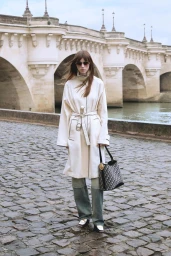 Model wearing a cream belted coat and dark sunglasses while holding a patterned tote bag on a cobblestone path beside an arched stone bridge.