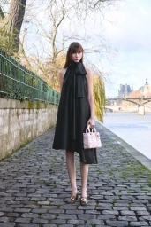 Model wearing a black sleeveless high-neck dress and carrying a pale pink handbag on a cobblestone riverside promenade.
