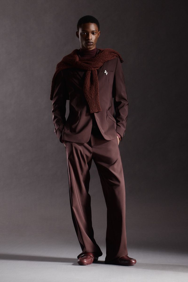 Man in brown suit and scarf standing against a dark grey backdrop in a studio setting, fashion portrait.