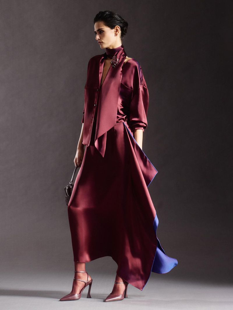 Fashion shot of a woman in a burgundy dress and matching high heels, holding a small purse, in a photo studio with a gray backdrop.