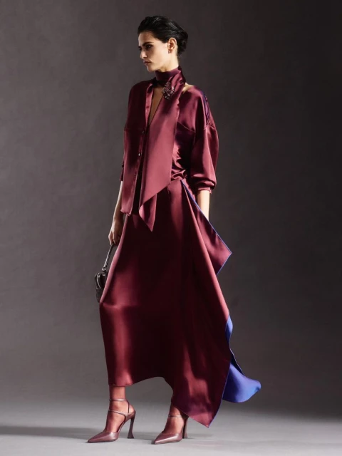 Fashion shot of a woman in a burgundy dress and matching high heels, holding a small purse, in a photo studio with a gray backdrop.
