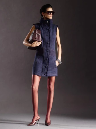 Model posing in a navy sleeveless high-collar mini dress with oversized sunglasses and a burgundy clutch bag against a grey backdrop.