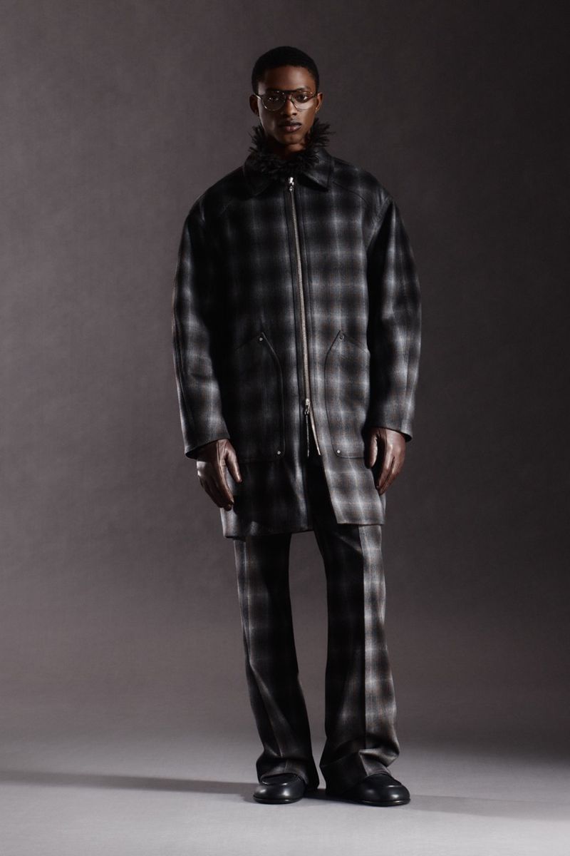 Full shot of a stylish Black man in a gray plaid coat with a fur collar and matching pants against a neutral gray backdrop.