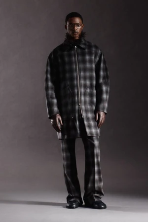 Full shot of a stylish Black man in a gray plaid coat with a fur collar and matching pants against a neutral gray backdrop.