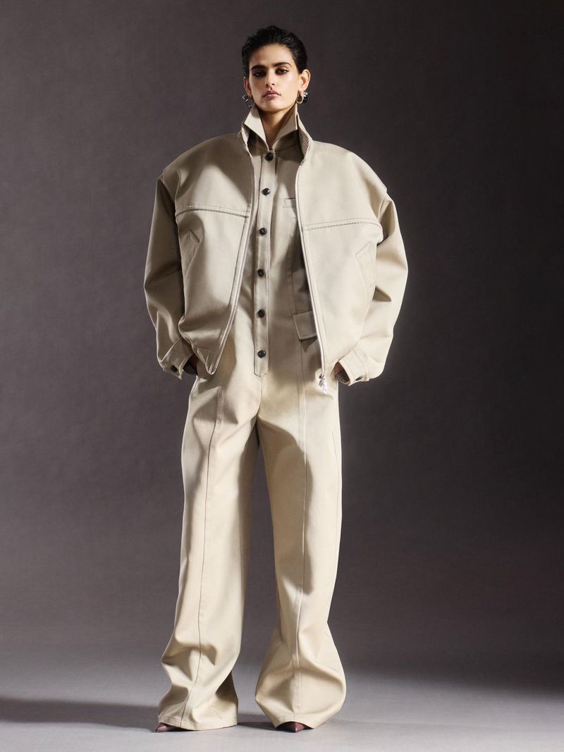 An adult fashion model stands in a photo studio wearing a light tan zipper jacket and matching jumpsuit with flared legs against a grey background.
