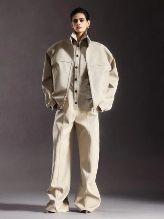 An adult fashion model stands in a photo studio wearing a light tan zipper jacket and matching jumpsuit with flared legs against a grey background.